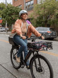 Woman riding the RadKick Belt Drive in MIdnight Blue down a city street. She is wearing an orange jacket & a helmet. The bike is equipped with a large front-mounted basket & rear-mounted hard shell locking box. 