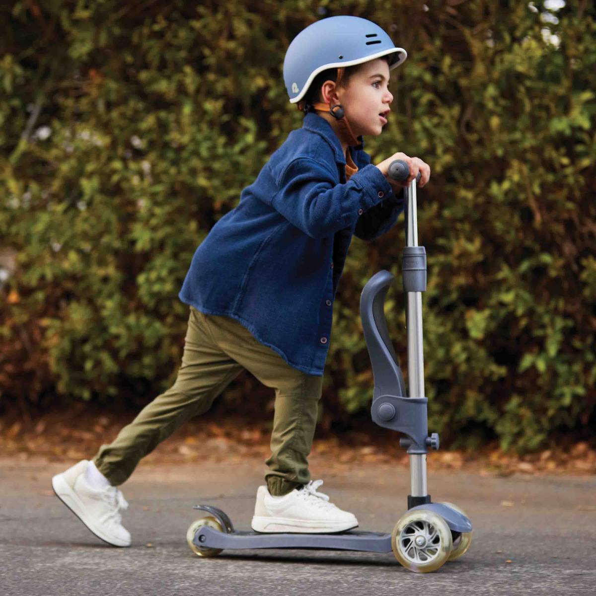 A child confidently rides a scooter down a quiet, tree-lined street while wearing a blue helmet and casual clothing.