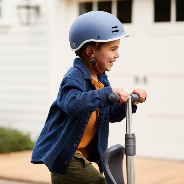 A child wearing a blue helmet and a navy sweater rides a scooter on a driveway, with a home in the background.
