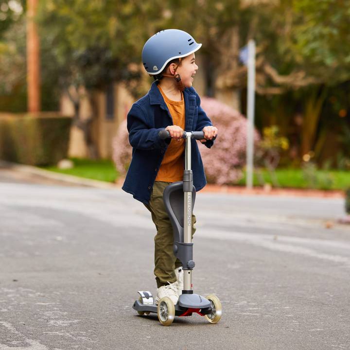 A child confidently rides a scooter down a quiet, tree-lined street while wearing a blue helmet and casual clothing.