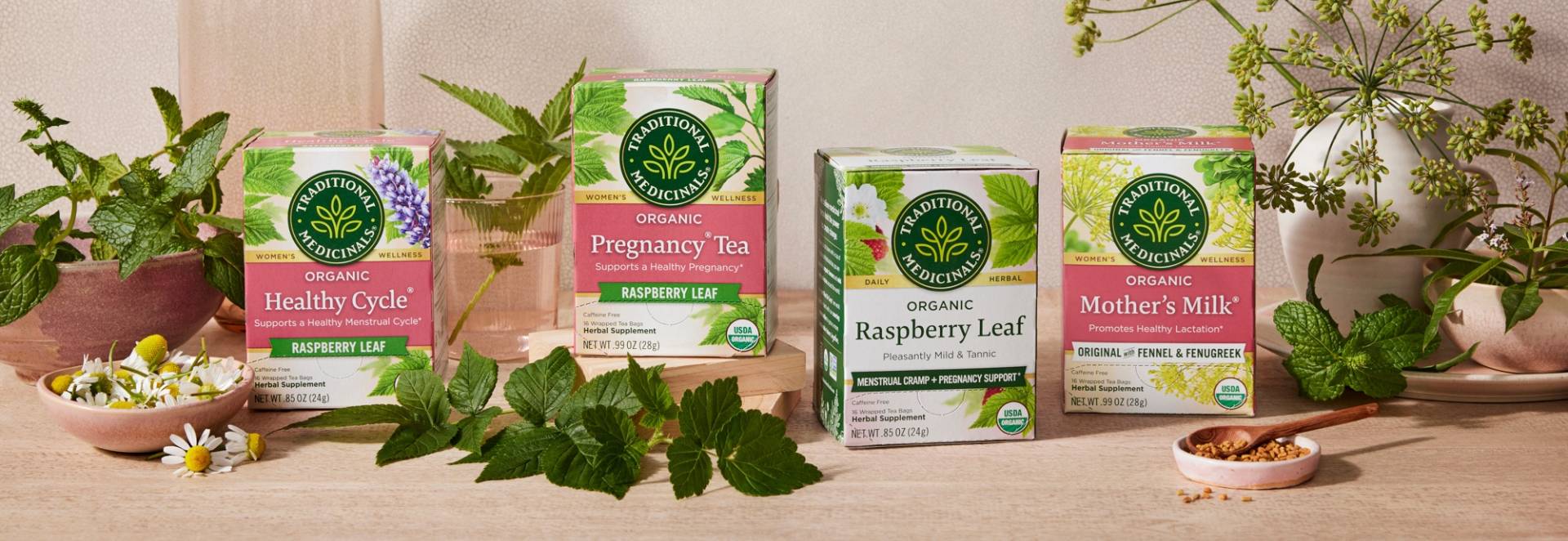 Boxes of teas including Healthy Cycle, Pregancy Tea, Raspberry Leaf, and Mother's Milk on a table with fresh herbs and flowers