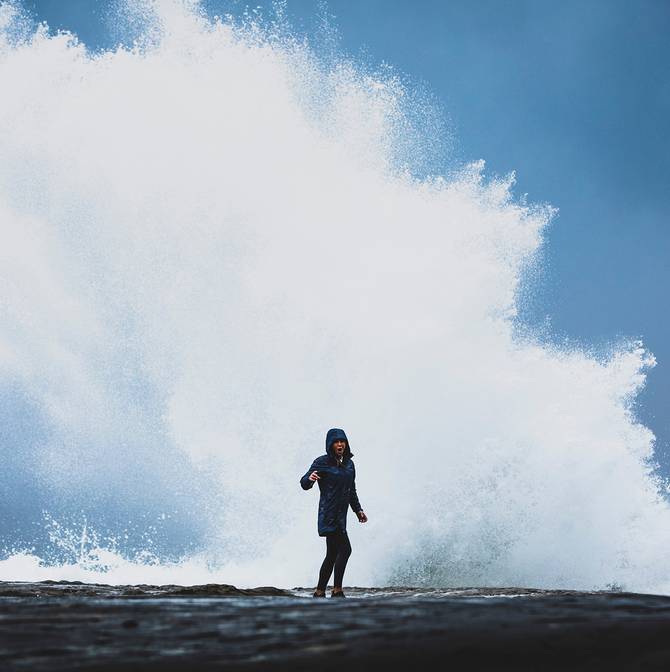 Women standing in front of wave crashing on rock ledge, she's wearing a long blue Aden rain jacket.