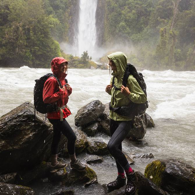 Women hiking by a waterfall wearing rain jackets and backpacks with their hoods up.