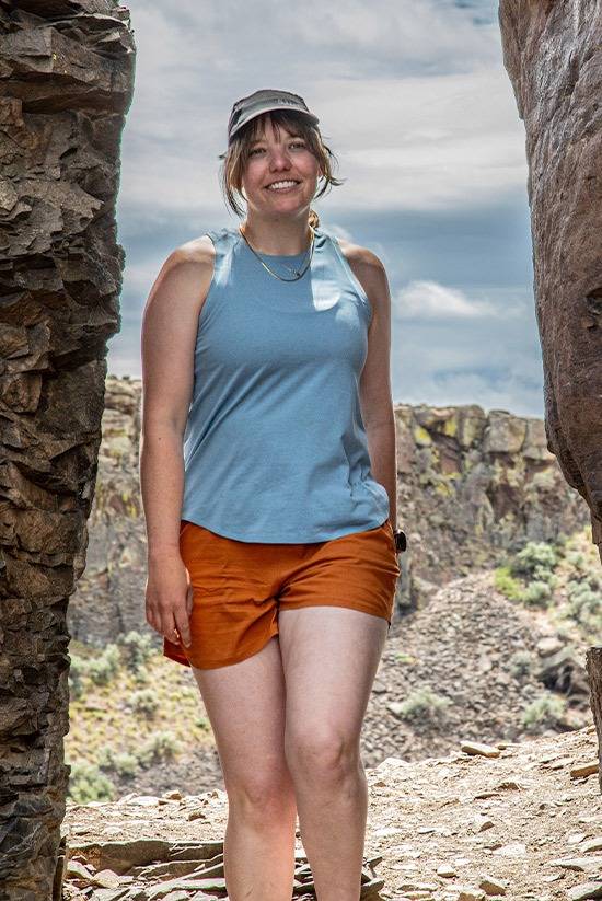 Woman standing between two rock walls with a high desert landscape in the background. Wearing the Big Sky Blue Feathers Tech-Tank and the Desert Clay OlallieLoop shorts. Smiling look at the camera.