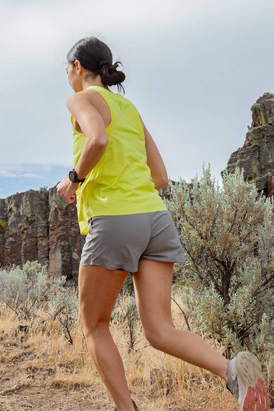 Woman wearing the Light Lime colored Feathers Tech-Tank and the Moonstone colored OlallieLoop Shorts while train running in a high-desert environment. Running away from the camera.