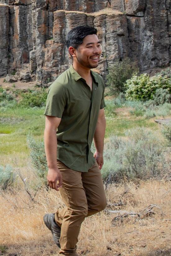 Man walking from left to right in a high desert landscape with rock formations in the background. He is wearing the Evergreen FirstSun Shirt and the Tobacco TarnGood Pants.