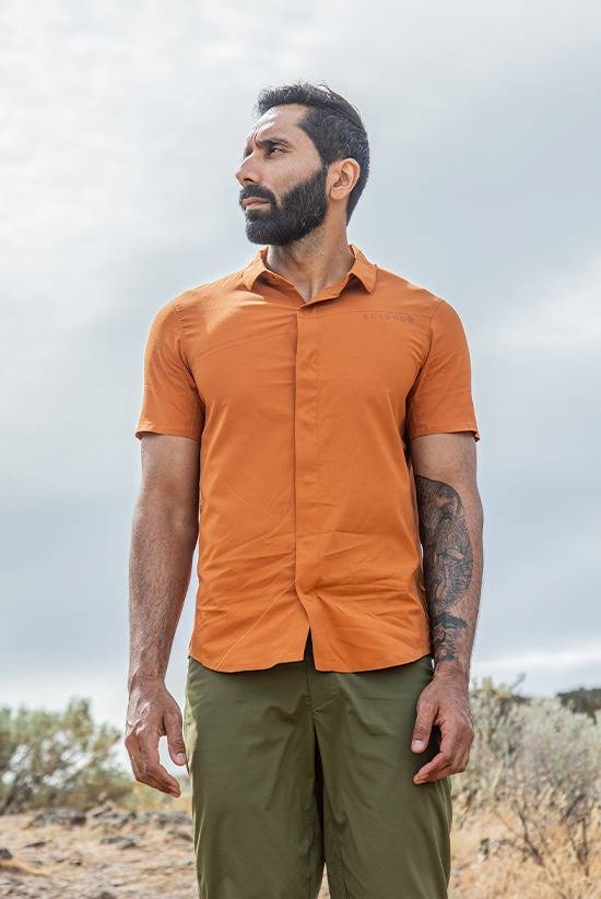 Man wearing the Desert Clay FirstSun Shirt and Evergreen TarnGood pant while looking off to the side with a cloudy sky background.