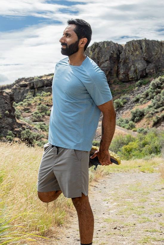 Man stretching on the trail while wearing the Moonstone colored TarnGood shorts and the Big Sky Blue SolarSwift Tech-T. Background is a high desert landscape with partially cloudy sky.