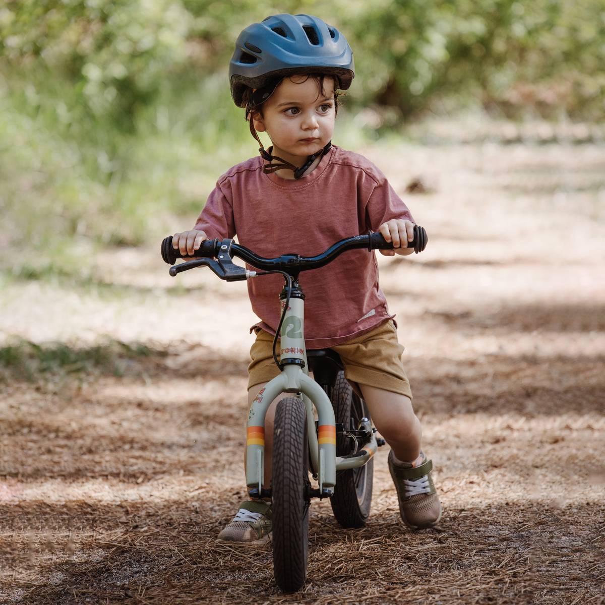 A young child wearing a helmet rides a Cub Plus 12" Kids' Balance Bike on a dirt path surrounded by greenery. The sun filters through the trees.