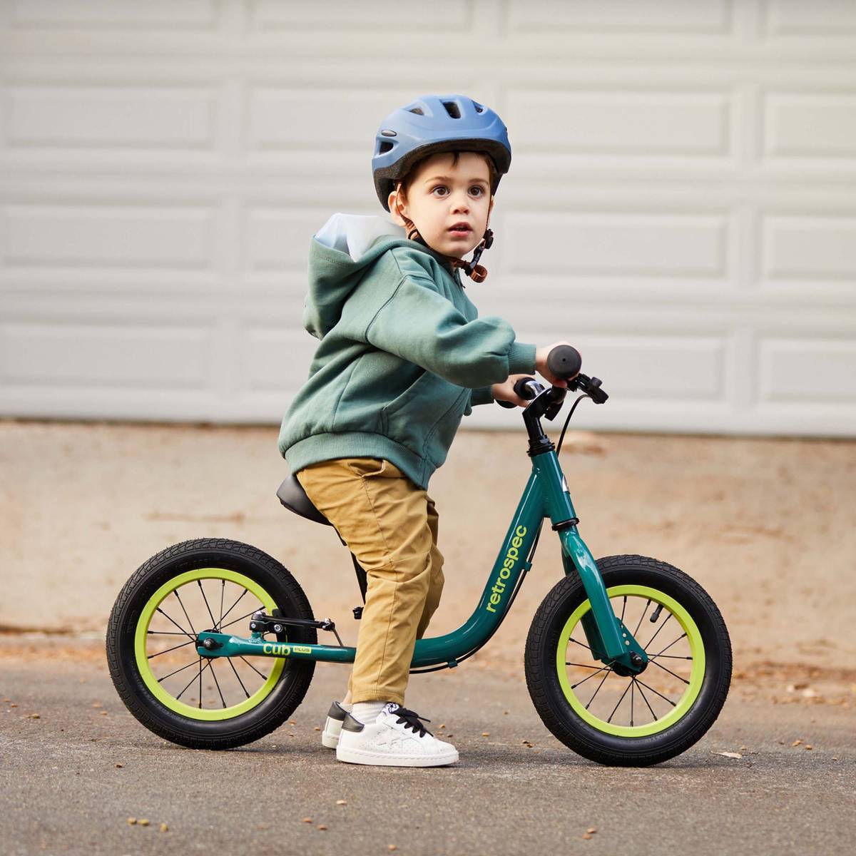 A child with a teal Cub Plus 12" Balance Bike wearing a blue helmet and a green hoodie, on a paved path.