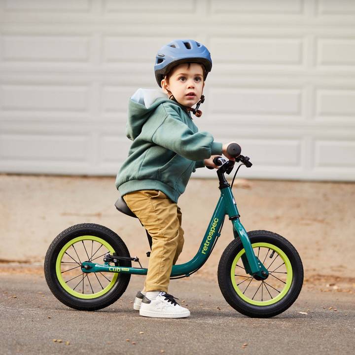 A child with a teal Cub Plus 12" Balance Bike wearing a blue helmet and a green hoodie, on a paved path.