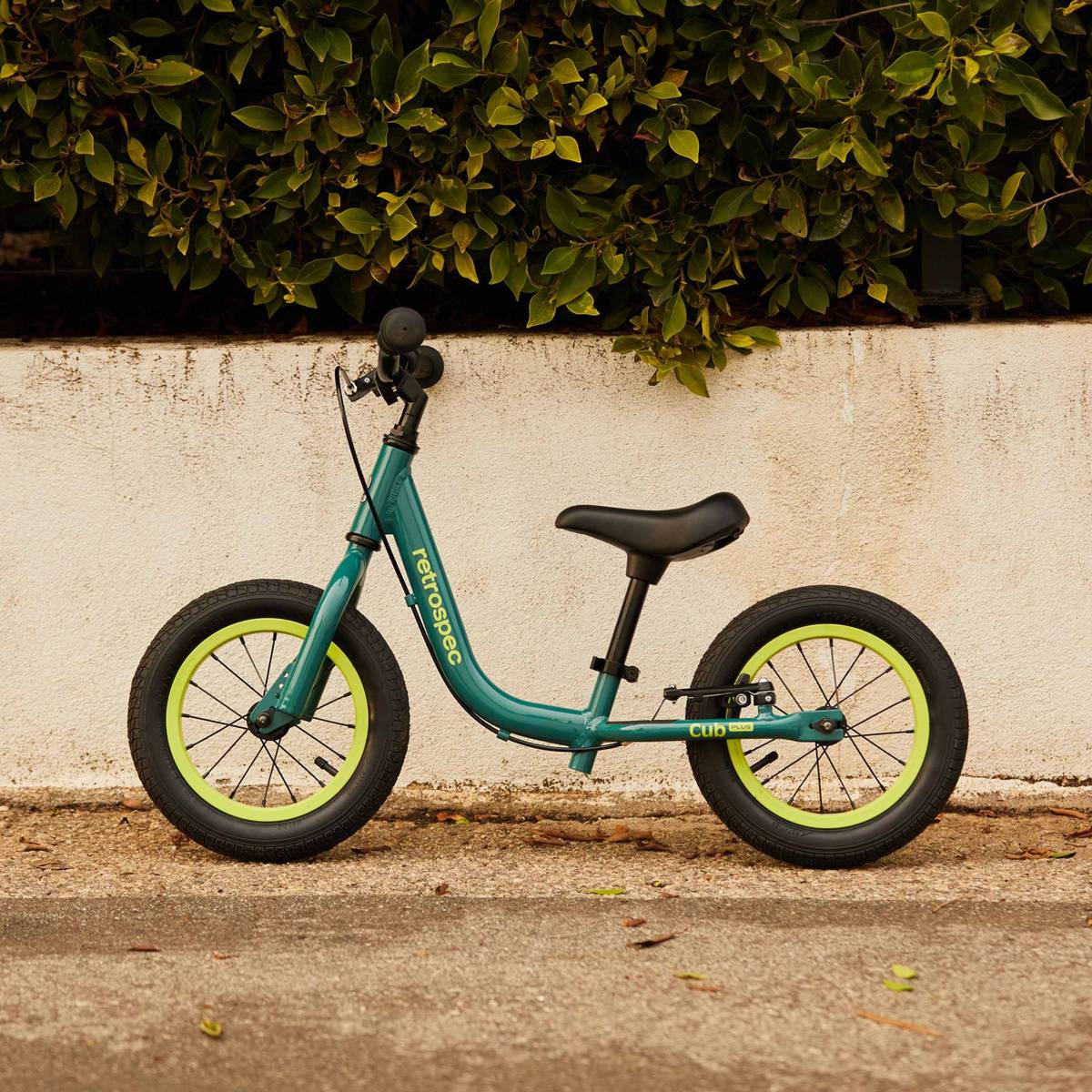 A teal Cub Plus 12" Balance Bike with black seat and lime green wheels, positioned against a white wall and foliage backdrop.