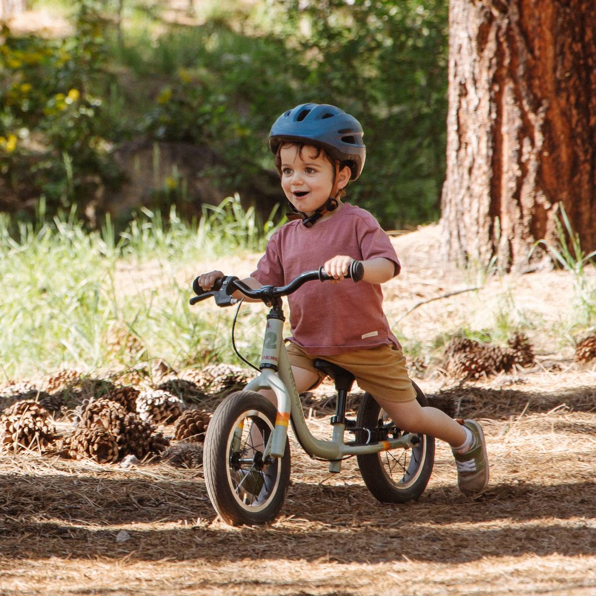 A child rides a Cub Plus 12" Kids' Balance Bike through a forested path, wearing a helmet and casual clothing, enjoying outdoor play in nature.