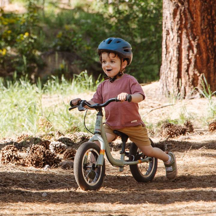 A child rides a Cub Plus 12" Kids' Balance Bike through a forested path, wearing a helmet and casual clothing, enjoying outdoor play in nature.