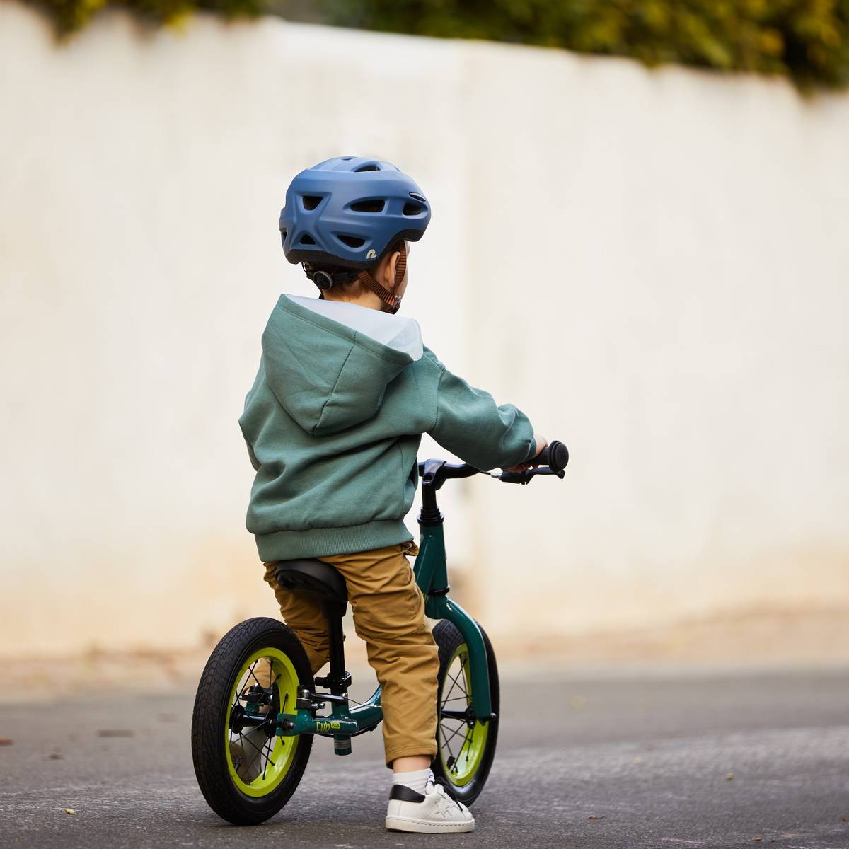 A child in a teal hoodie and helmet rides a green Cub Plus 12" Balance Bike on a quiet street.