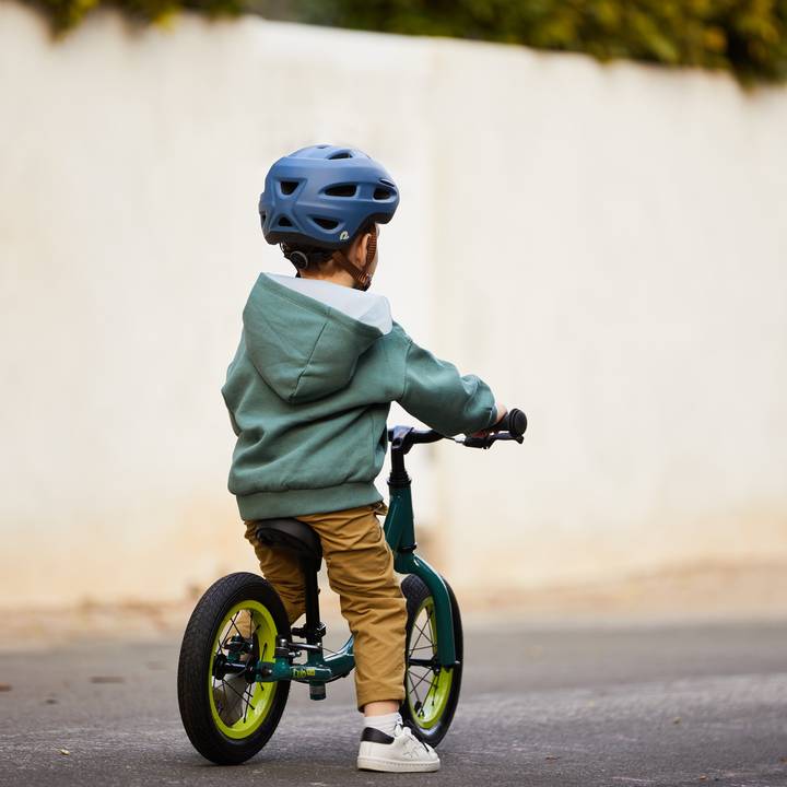 A child in a teal hoodie and helmet rides a green Cub Plus 12" Balance Bike on a quiet street.