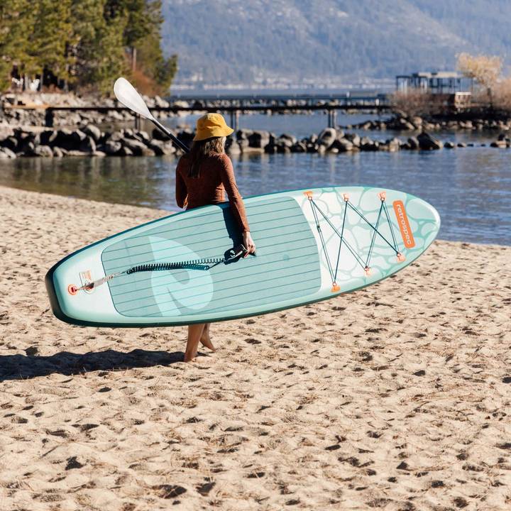 Person in a yellow hat carrying a paddleboard and paddle on a sandy beach near calm water with trees and mountains in the background.