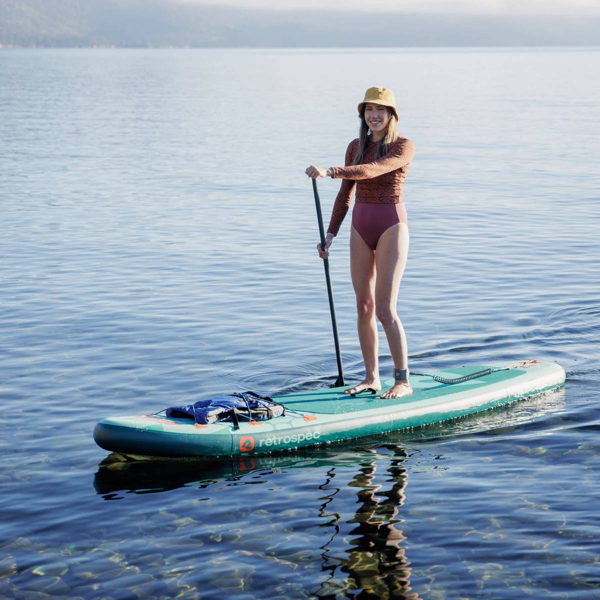 Person paddleboarding on calm water, wearing a hat and long-sleeve top, with a bag on the board under clear daylight.