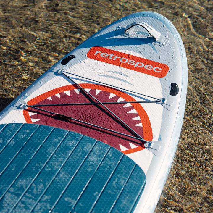 A Weekender Nano Inflatable Stand-Up paddleboard for kids with a shark graphic lies on clear water, featuring the "retrospec" logo prominently.