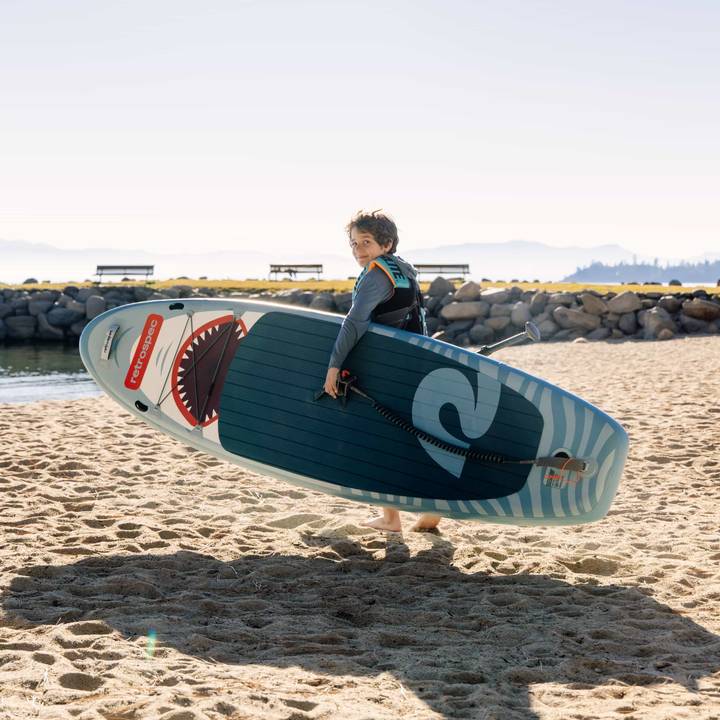 A child carrying a Weekender Nano Inflatable Paddleboard with a shark design walks on sandy shores near a tranquil lake, framed by distant mountains.