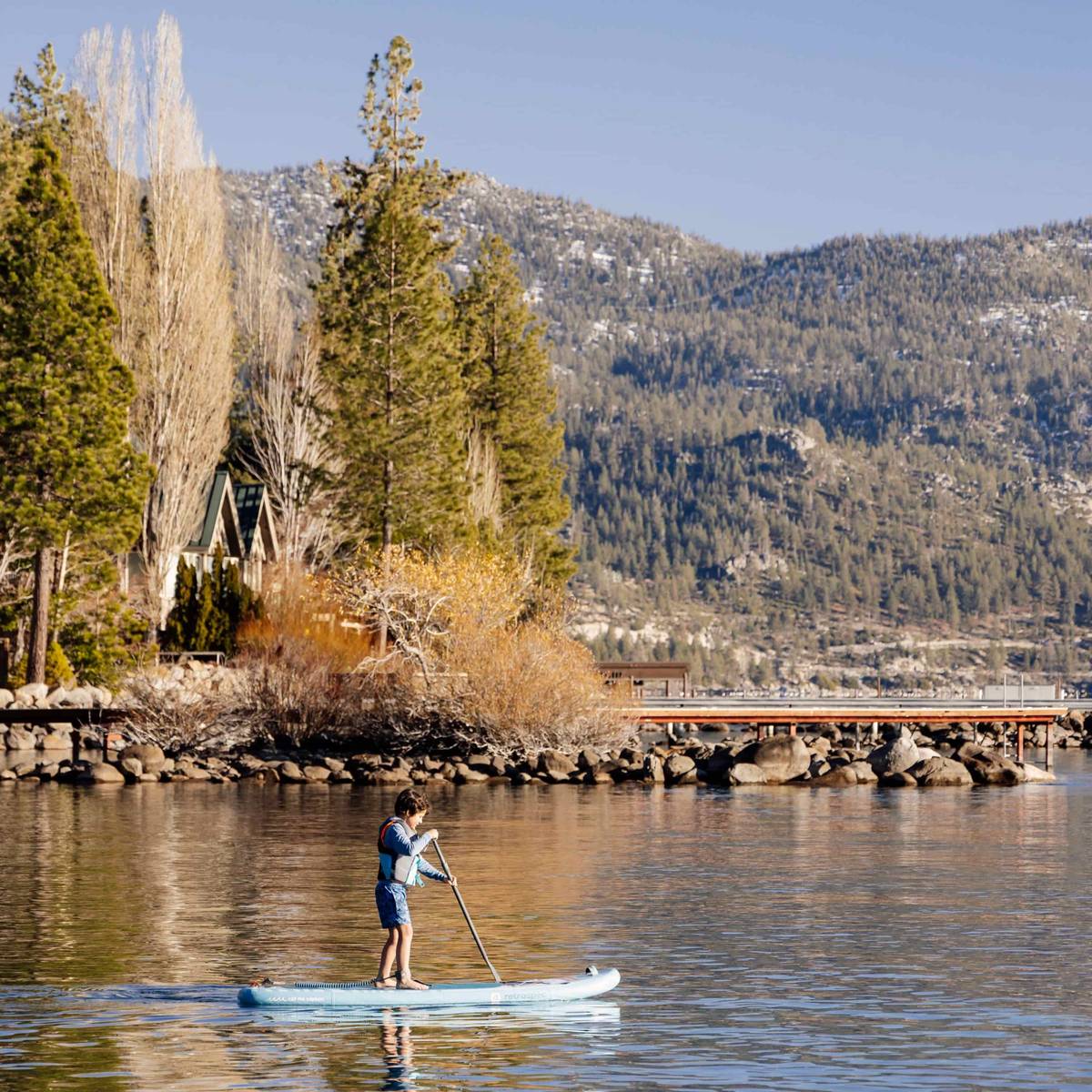 A child paddleboarding on calm water, surrounded by tall trees and mountains under a clear blue sky.