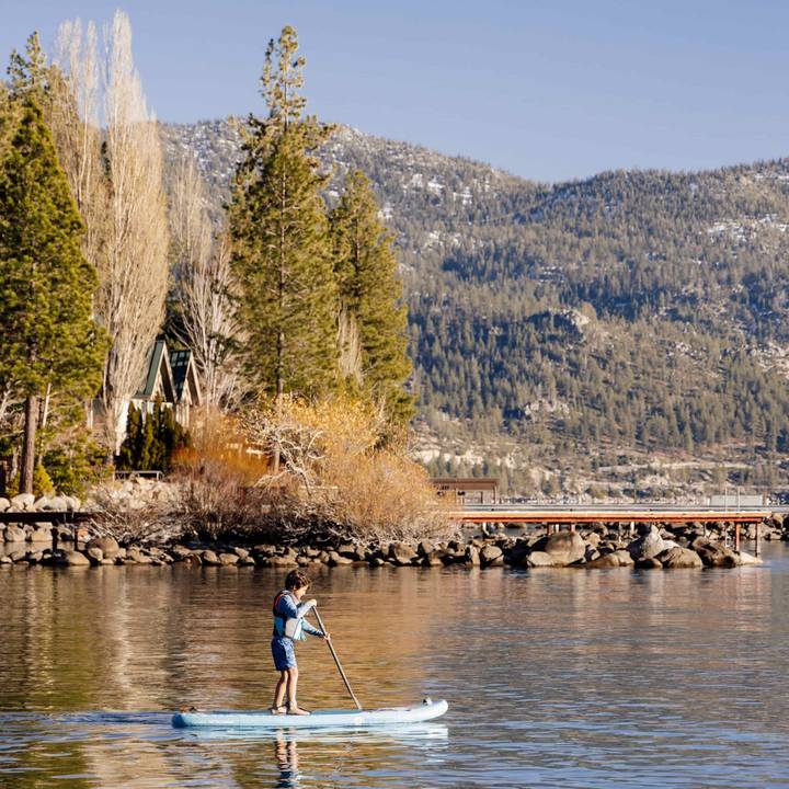 A child paddleboarding on calm water, surrounded by tall trees and mountains under a clear blue sky.