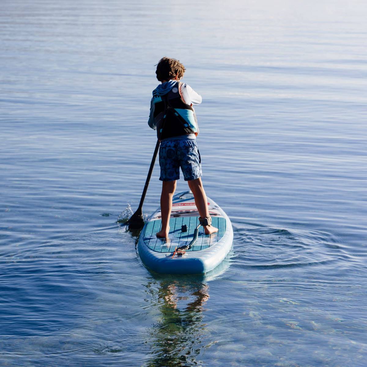 A child stands on a kids inflatable paddleboard, paddling through calm waters with a serene background, enjoying a sunny day outdoors.