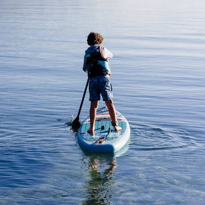 A child stands on a kids inflatable paddleboard, paddling through calm waters with a serene background, enjoying a sunny day outdoors.