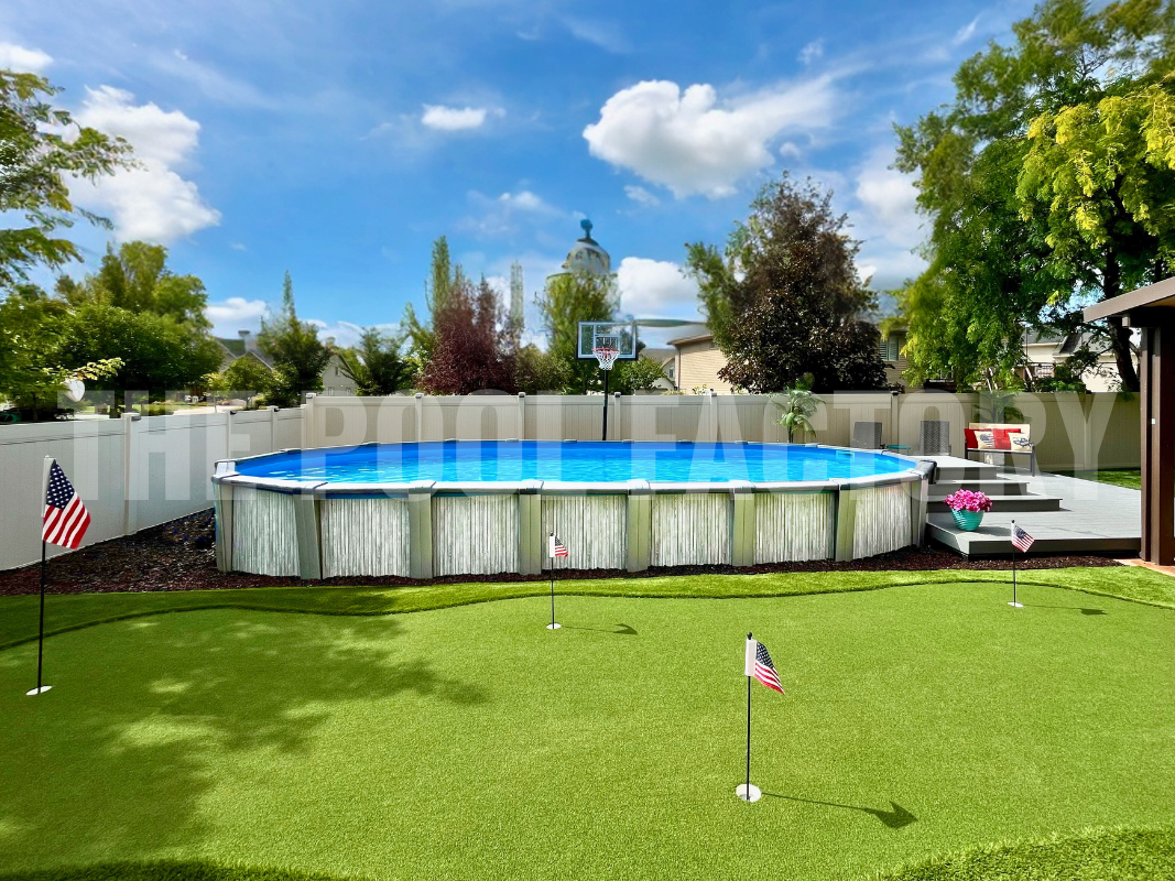 Oval swimming pool with deck, putting green, and trees under bright skies