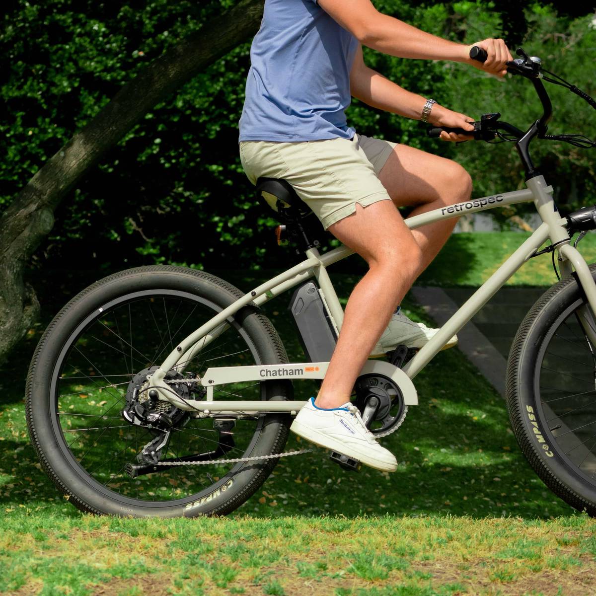 Person wearing a blue shirt and khaki shorts rides a Chatham Rev Beach Cruiser Electric Bike on short green grass.