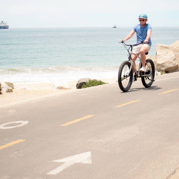 Person wearing a blue helmet and shirt rides a Chatham Rev Beach Cruiser Electric Bike on a bike path with the ocean in the background.
