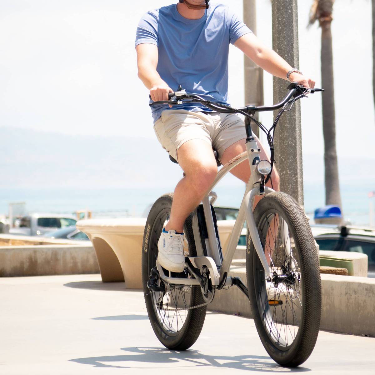 Person wearing a blue shirt and khaki shorts rides a Chatham Rev Beach Cruiser Electric Bike on a bike path with the ocean in the background.