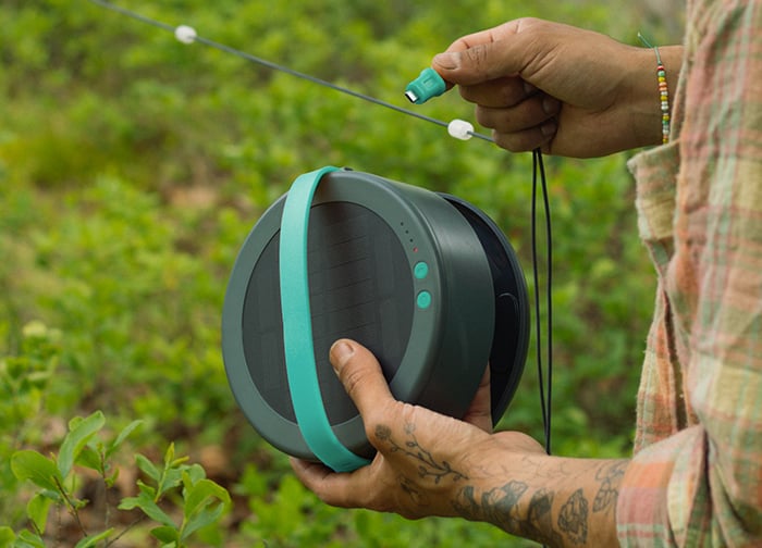 A person holds a portable speaker with a green strap, preparing to connect it to a power source in a lush outdoor setting.