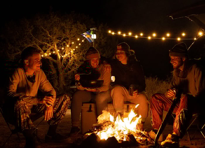 A group of four friends gathered around a campfire at night, enjoying food and drinks, with string lights and a flag in the background.