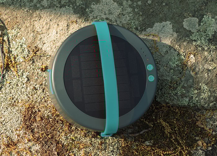 A round, portable speaker with a solar panel on top, featuring a teal strap, resting on a rocky, moss-covered surface.