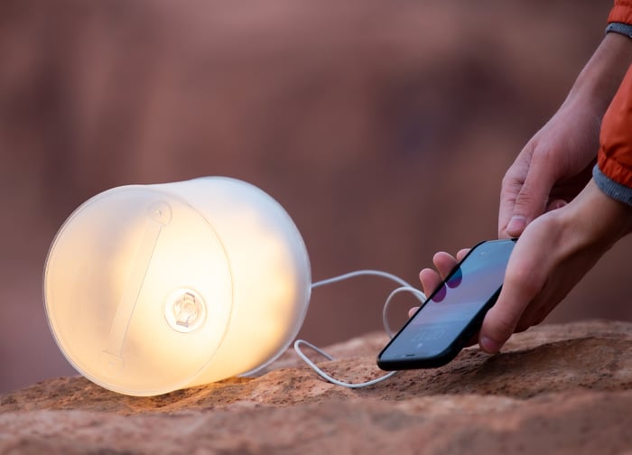 A person is charging a smartphone using a portable, glowing lamp on a rocky surface.