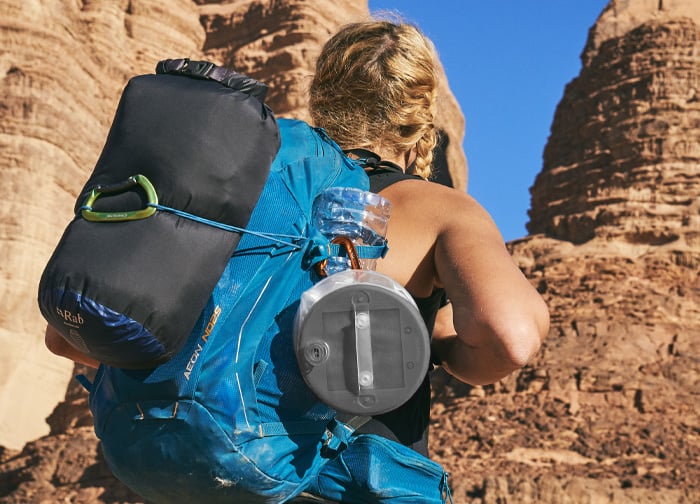 A person with a blue backpack and a sleeping bag is hiking in a rocky landscape under a clear blue sky.