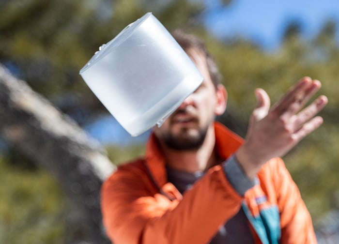 A person in an orange jacket is tossing a translucent container in the air against a blurred natural background.