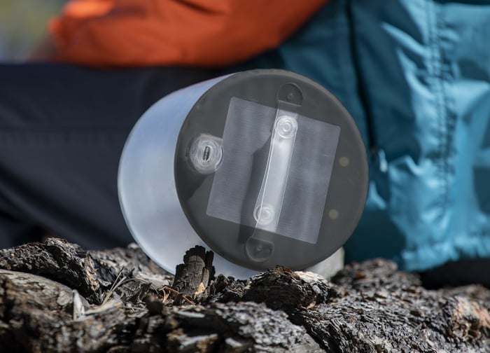 A transparent container with a solar panel sits on a log, with a person in an orange jacket and teal coat in the background.