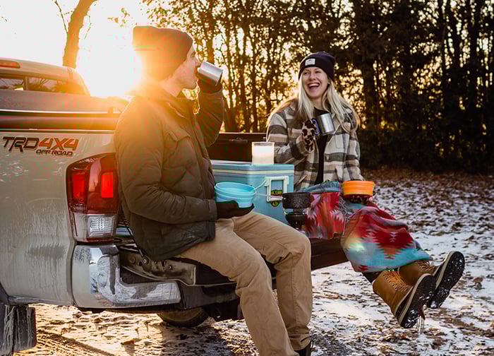 A couple sits on the tailgate of a truck, enjoying drinks and snacks in a snowy setting, with sunlight shining through trees.