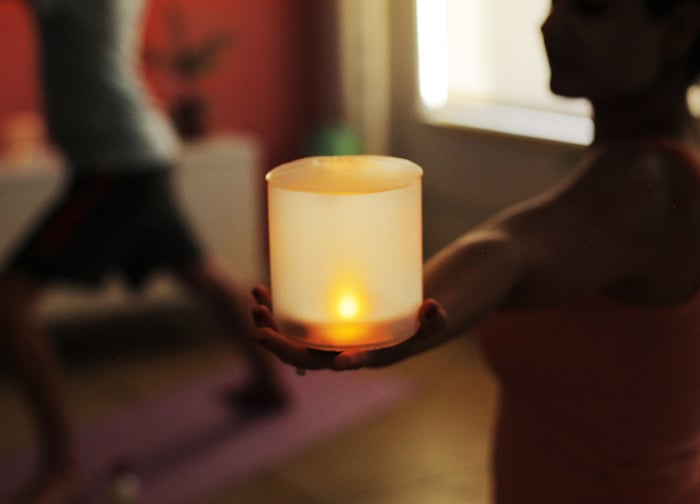 A person holds a glowing candle in a serene setting, with a blurred figure practicing yoga in the background.