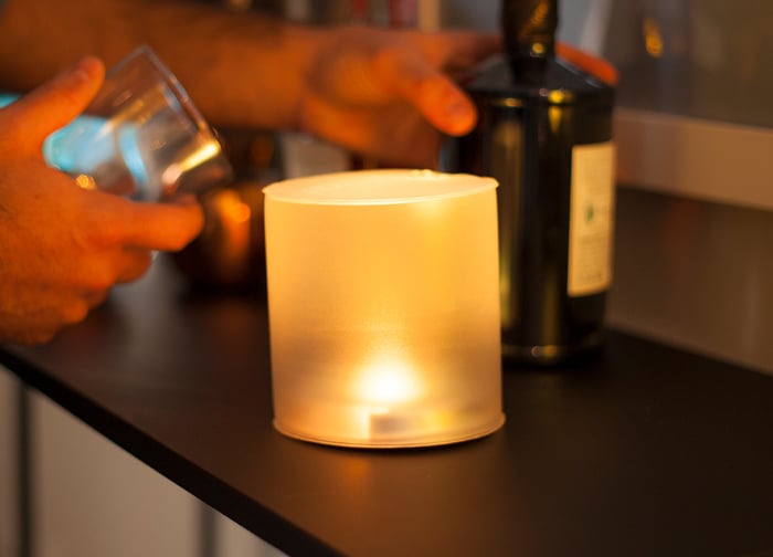 A softly glowing candle sits on a dark table, with a person holding a glass and a bottle nearby.