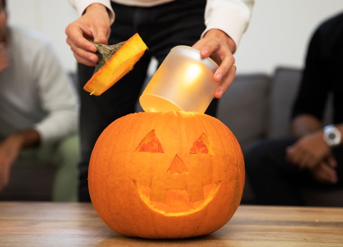 A person is placing a candle inside a carved pumpkin with a smiling face, while others watch in the background.