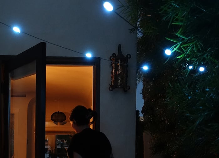 A person stands at an open door, illuminated by warm light, with string lights glowing nearby in a dimly lit outdoor setting.