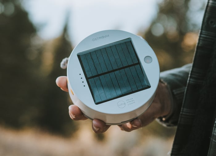 A person holds a round solar-powered light with a grid pattern on top, set against a blurred outdoor background.