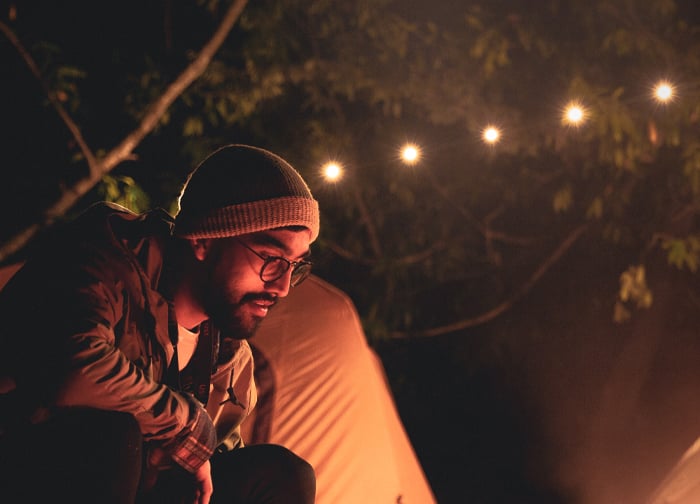 A man in a beanie sits near a campfire, surrounded by tents and string lights in a forested area.