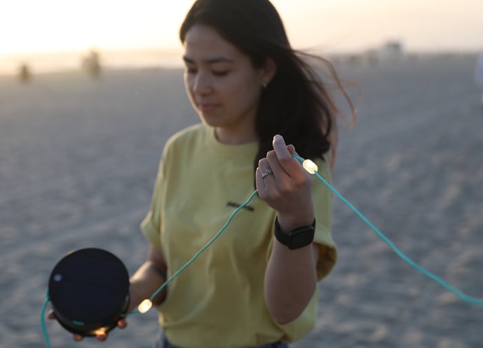 A person in a yellow shirt holds a solar panel and string lights on a beach during sunset.