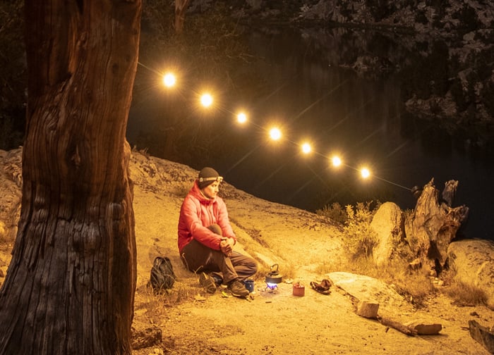 A person sits by a lake at night, surrounded by warm string lights, with camping gear nearby. The scene is serene and illuminated.