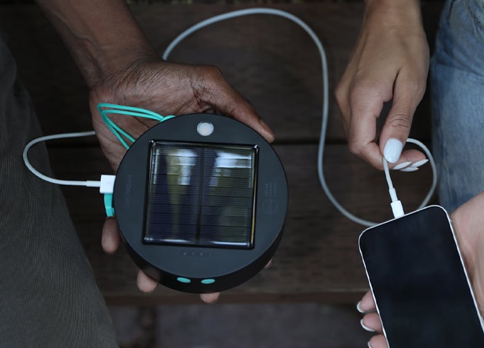 A solar charger is being used to charge a smartphone, with hands holding the charger and phone.