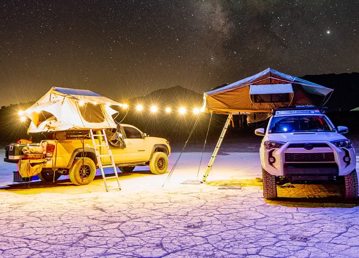 Two vehicles with rooftop tents are set up under a starry night sky, illuminated by string lights on a dry, cracked landscape.